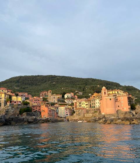 Pastel houses cling to a hillside village above the Ligurian Sea under soft evening light.