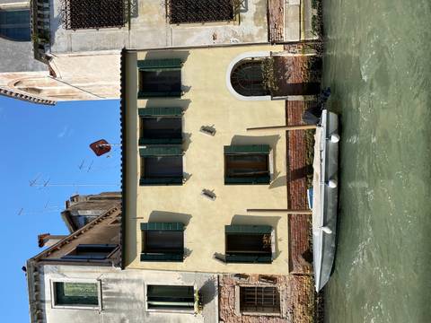 Small yellow house with green shutters sits beside a narrow Venetian canal with a moored dinghy.