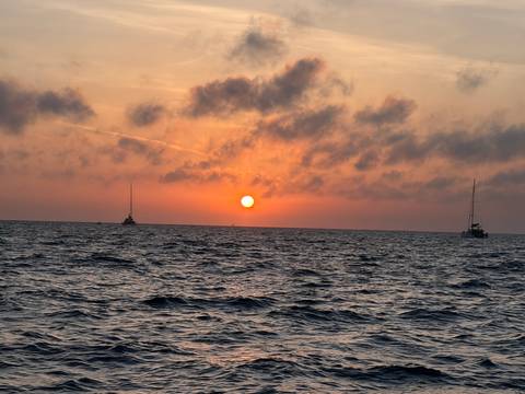 Fiery orange sun sets over the Aegean Sea with sailboats silhouetted on the horizon.