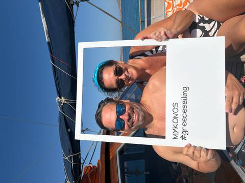 Smiling couple on a yacht holds a white photo frame labelled 'MYKONOS #greecesailing' against blue sky.