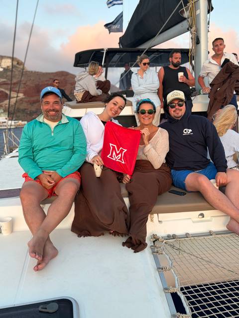 Group bundled in blankets on a catamaran deck at sunset proudly holds a red flag.