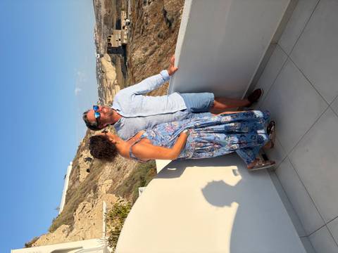 Couple pose on a whitewashed balcony with Santorini’s cliffside village behind in late afternoon light.