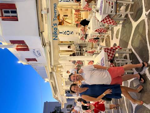 Couple stands outside the brightly lit Taverna Nikos on a Mykonos evening.