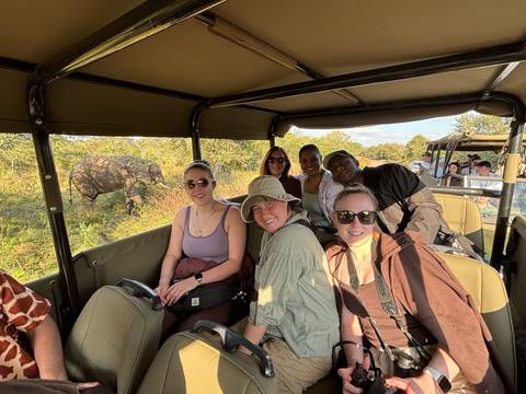 Safari-goers in open jeep smiling while an elephant browses in the bush nearby.