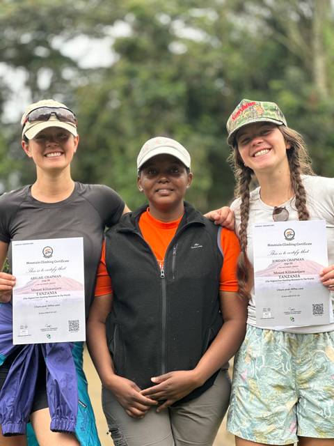 Three climbers proudly display Mount Kilimanjaro summit certificates, smiling for the camera outdoors.