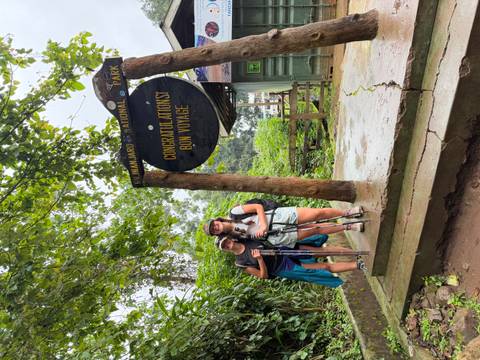 Two trekkers with poles pose beneath a wooden Kilimanjaro National Park gate reading 'Congratulations! Bon Voyage'.
