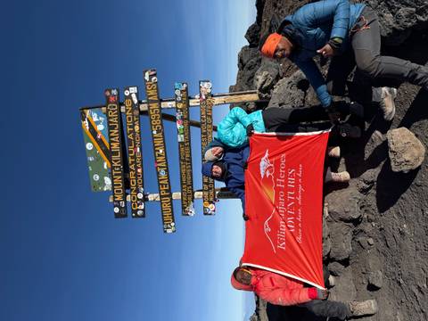 Climbers at Uhuru Peak sign on Mount Kilimanjaro holding a red expedition flag under clear skies.