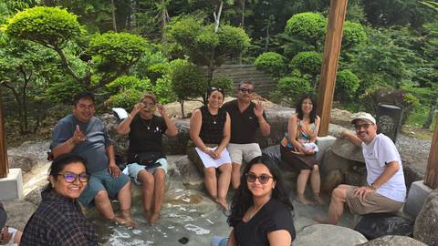 Group of travellers soaking feet in an outdoor hot-spring foot bath surrounded by lush garden.