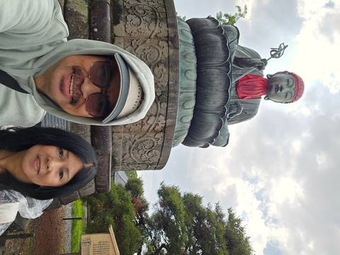 Selfie of couple in front of large Jizo statue atop a stone pedestal with cloudy sky.