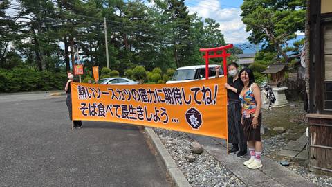 Travellers hold a long orange banner with Japanese text outside a rural roadside eatery near Mount Fuji area.