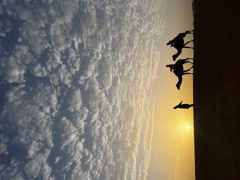 Silhouettes of guide leading two camel riders across a desert ridge at sunrise with dramatic cloudscape.