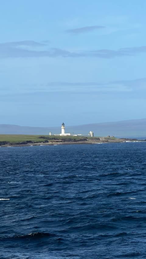 White lighthouse on a grassy headland surrounded by blue sea with hazy hills in the distance