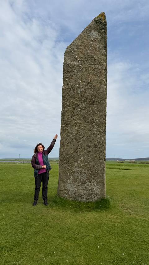 Woman pointing up beside a tall monolithic standing stone on grassy plain