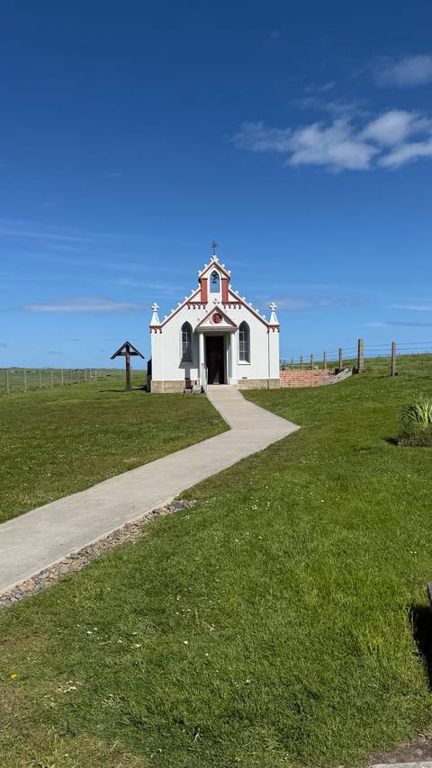 Small white Italian Chapel with red trim standing alone in a grassy field under a bright blue sky