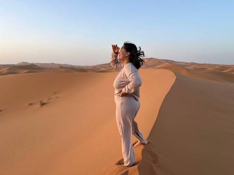 Woman in light loungewear stands on a high dune shading her eyes while gazing across endless sand waves