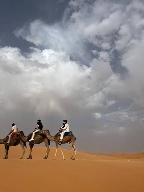 Three travellers ride camels beneath dramatic storm clouds over the desert