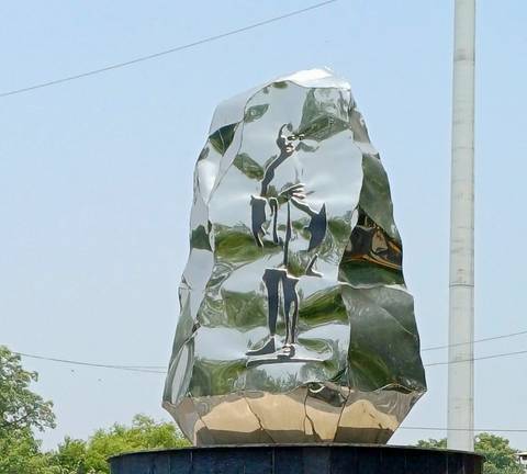 Abstract polished-steel sculpture gleaming under daylight with a lamppost in the background.