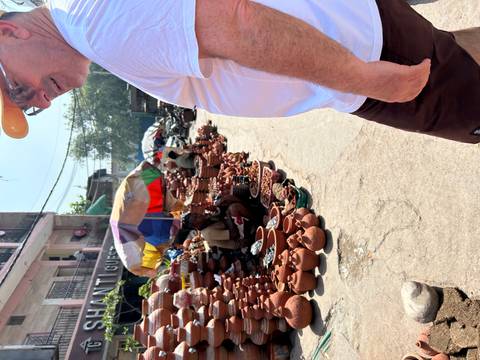 Outdoor pottery market with terracotta wares piled high under a colorful umbrella and shoppers passing by.