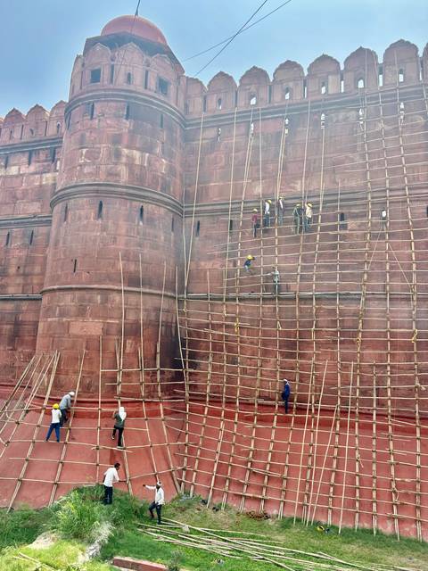 Construction workers scaling bamboo scaffolding on a massive red fort wall during restoration.