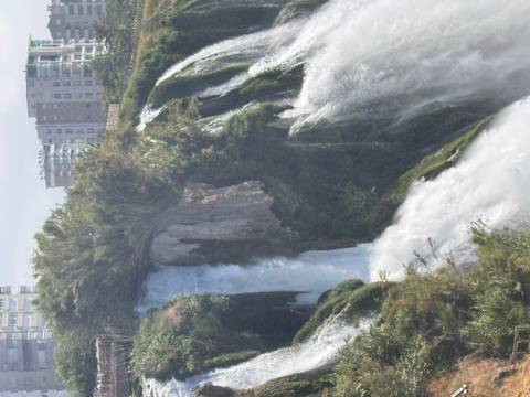 A powerful urban waterfall cascades over a cliff framed by greenery with modern apartment blocks visible on the skyline.