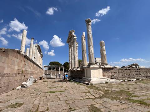 Sun-lit marble columns and ruins of an ancient temple dominate a hilltop archaeological site under a vivid blue sky, with two tourists walking along the stone paving.