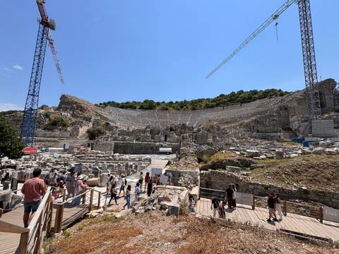 A vast stone amphitheatre under restoration, filled with visitors exploring the ancient seating tiers as cranes loom overhead.