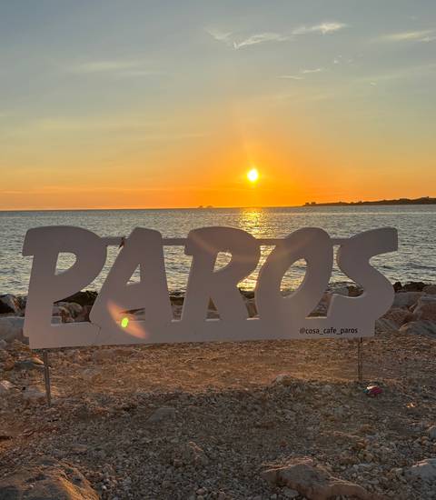 Silhouette of a large PAROS sign on a rocky shore at sunset with orange sky reflecting on the sea.