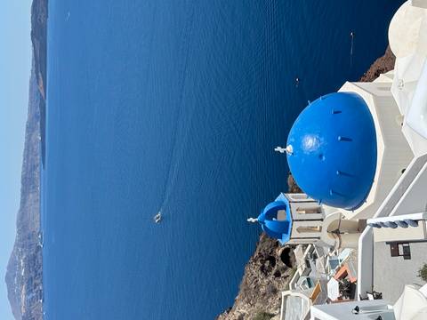 Iconic white church with bright blue dome perched on cliffs above the deep-blue Aegean Sea with a small boat below.