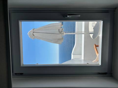 View through a white-framed window to a balcony umbrella and endless blue sea under cloudless sky.