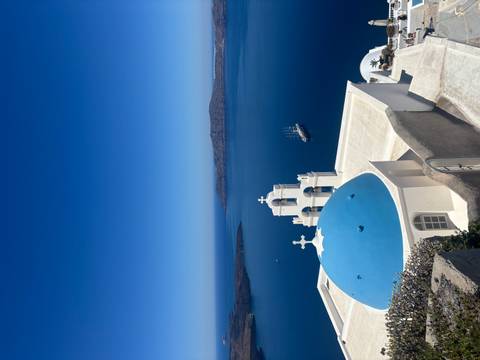 Classic white church with blue dome overlooking Santorini caldera and a tall-masted ship below under brilliant skies.