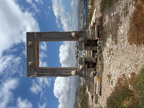 Ancient marble gateway (Portara) stands on a hilltop framing the town and harbor beneath a partly cloudy sky.