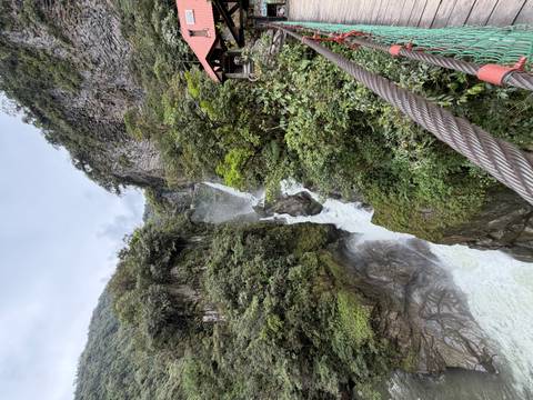 A powerful waterfall cascades through a lush green canyon viewed from a suspension bridge cable.