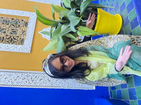A woman draped in colorful scarf smiles in front of vivid yellow and blue walls and patterned tiles at Jardin Majorelle.
