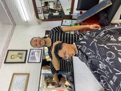 Barber smiles while giving a haircut inside a small barbershop decorated with photos and mirrors.