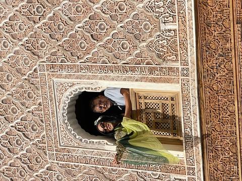 Travelers lean on an elaborately carved window balcony in a traditional Moroccan madrasa façade.