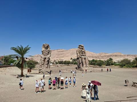 Tourists walk around the massive Colossi of Memnon statues under a clear blue sky.