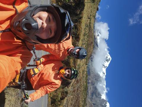 Three mountain bikers in orange jackets pose for a selfie with a snow-capped Andean peak behind.