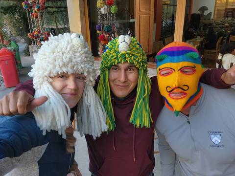 Three friends wear colorful traditional knit masks and smile at the camera in a busy market street.