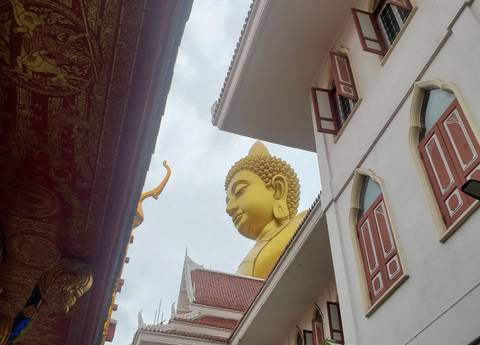 Upward perspective of a large golden Buddha head framed by temple buildings under a cloudy sky.