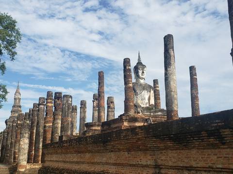 Ancient brick pillars and a seated Buddha statue at a historic temple ruin against a blue sky.