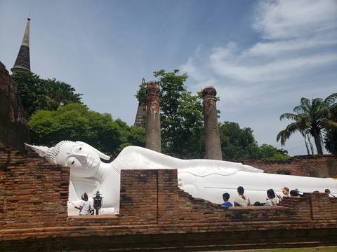Large white reclining Buddha statue lying amid brick ruins with visitors observing nearby.