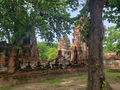 Shaded temple ruins with headless Buddha statues and red-brick chedis surrounded by leafy trees.