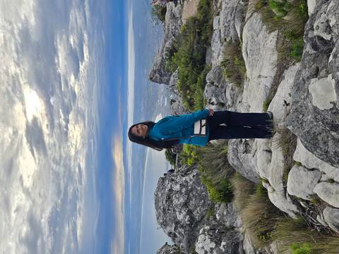 Woman poses on rocky outcrop of Table Mountain with dramatic clouds and Cape Town coastline below.