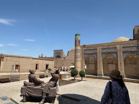 Historic courtyard in Khiva with brick madrasas, small minaret, statues and visitors under blue sky.