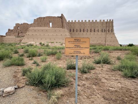 Earthen fortress ruins with an information sign and sparse desert vegetation under a cloudy sky.