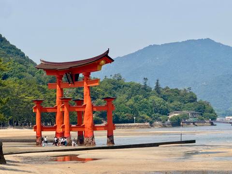 Large iconic red torii gate stands on tidal flats with lush green hills and blue mountains behind; small visitors gather at the base at low tide.