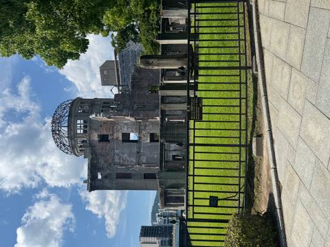 The ruined Atomic Bomb Dome rises behind an iron fence under a bright sky with patches of cloud and fresh green lawn in front.