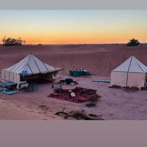 Small desert camp with two white canvas tents, rugs, and low tables set against orange dunes at sunset.