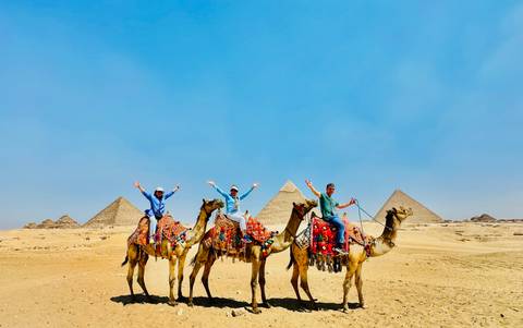 Riders on richly decorated camels raise their arms in front of the pyramids on the Giza Plateau under a blue sky.