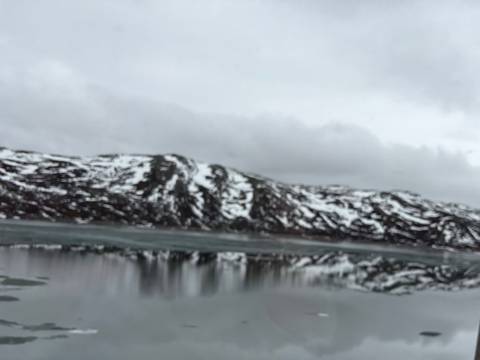 Blurry view of snow-covered mountains reflected in a still lake beneath grey clouds.